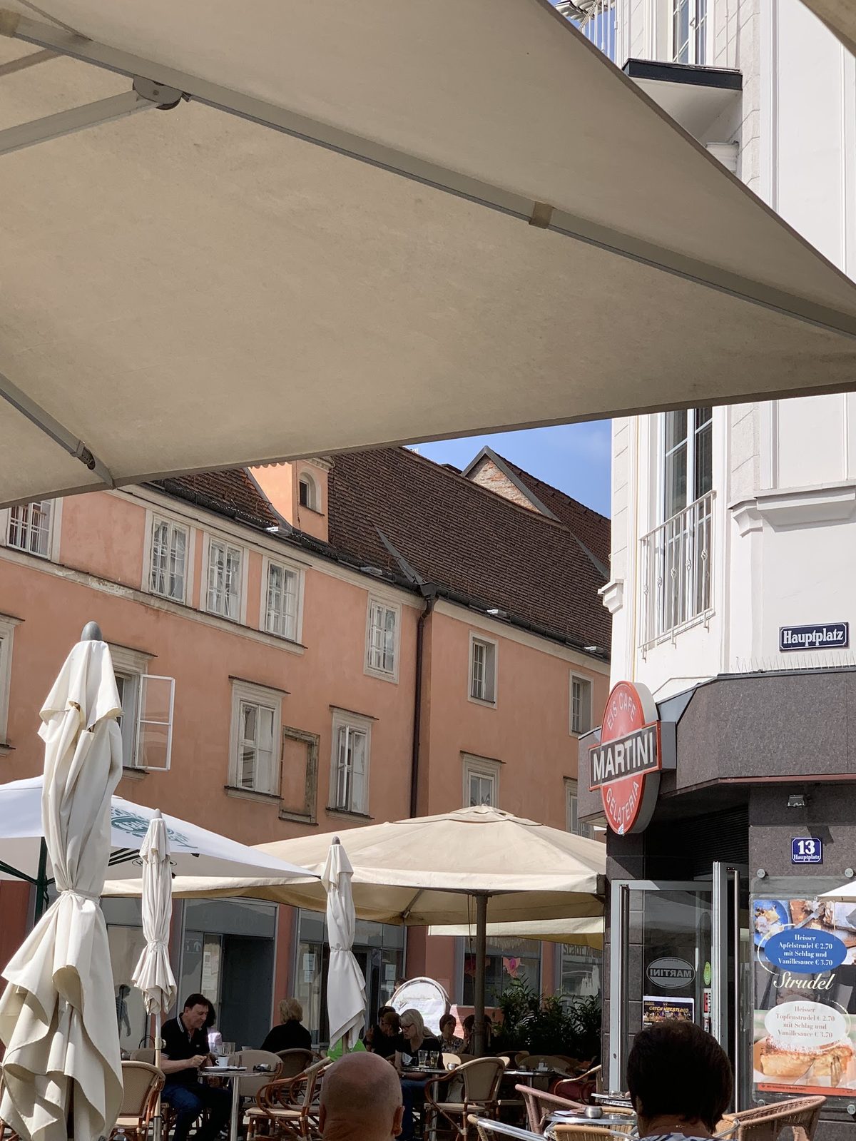 Gäste sitzen im Schanigarten unter einem großen Sonnenschirm vor dem Haus